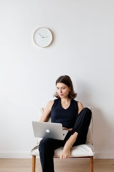 A woman sitting on a chair, using a laptop for remote work at home, with a clock on a white wall.