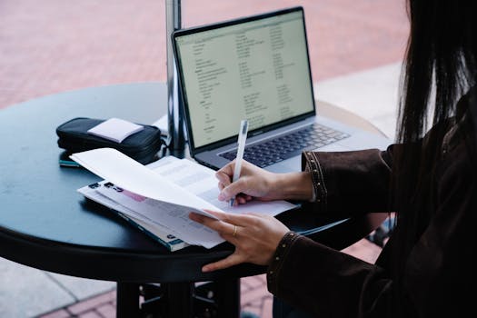 A woman writes on papers at an outdoor cafe table with an open laptop.