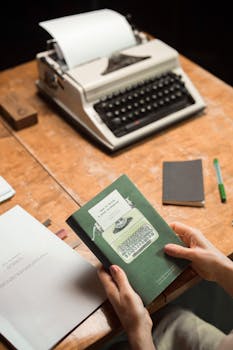 Hands hold a vintage book in front of an old typewriter on a wooden desk.
