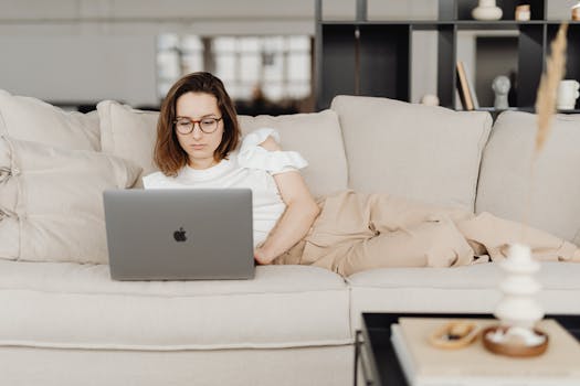 Woman wearing glasses relaxing on a beige couch while using a laptop in a modern living room.